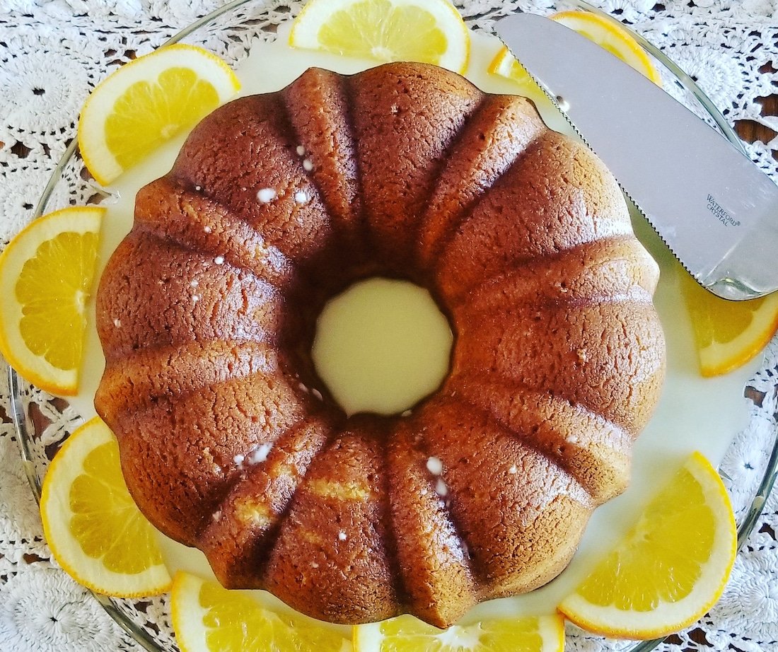 A glazed bundt cake sits on a glass plate surrounded by orange slices, with a knife placed next to it on a lacy white tablecloth.