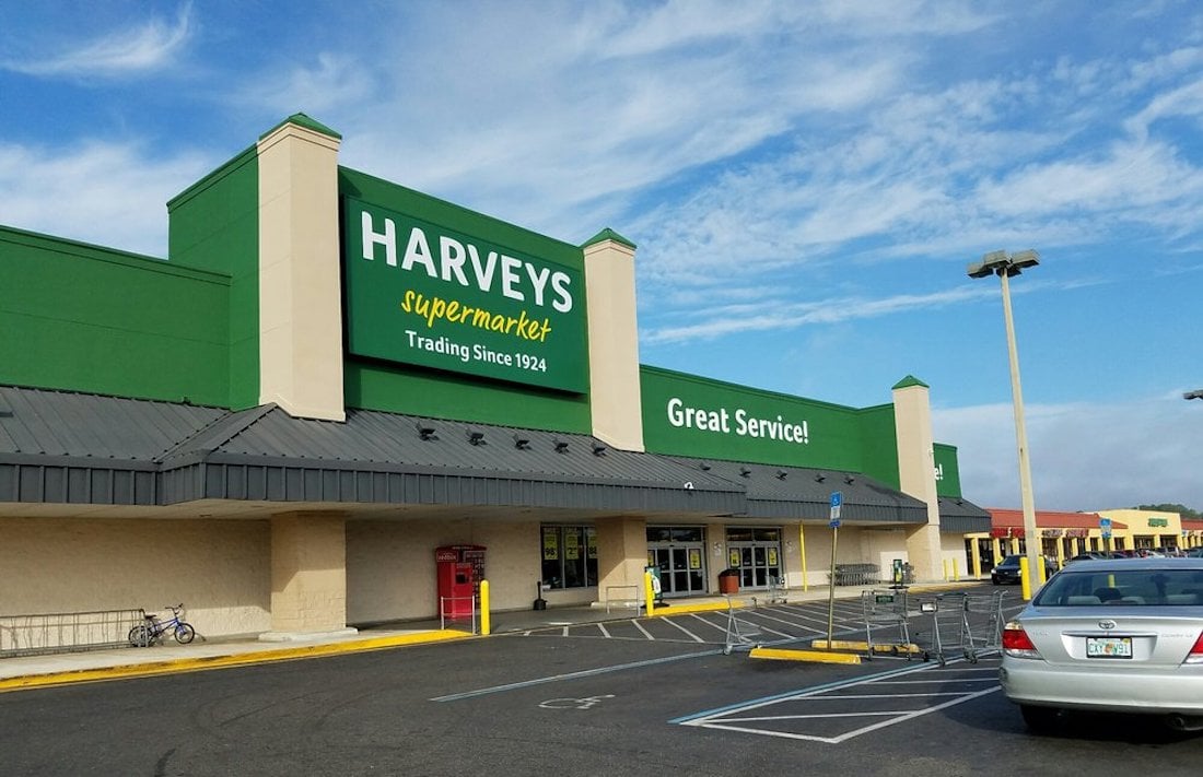 Harveys Supermarket storefront with green signage, visible parking lot, a few cars, bicycles, and shopping carts outside. The sign says "Great Service!" and the sky is partly cloudy.