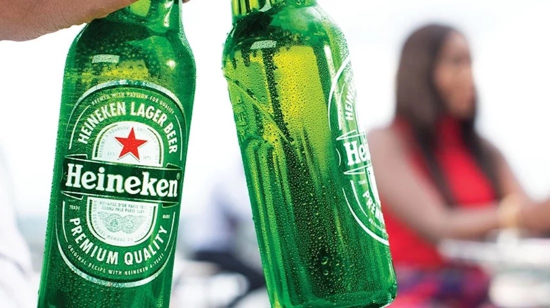 Two hands hold up green Heineken beer bottles in a toast, with condensation visible on the bottles. In the blurred background, a person sits at a table outdoors.