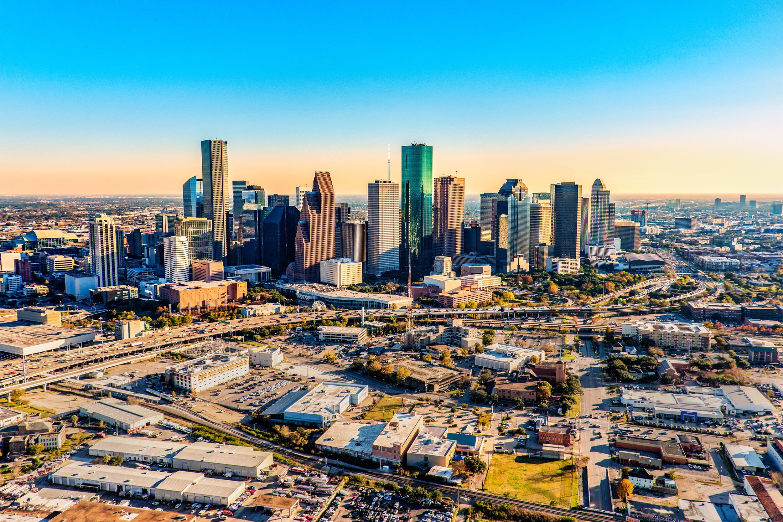 Aerial view of downtown Houston, Texas, featuring tall skyscrapers, highways, and surrounding industrial and residential areas under a clear blue sky.