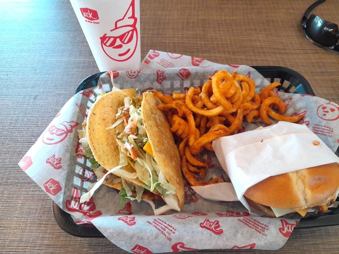 A fast food meal tray with two hard shell tacos, curly fries, a wrapped sandwich, and a large drink cup, all on branded paper from Jack in the Box, on a brown table.