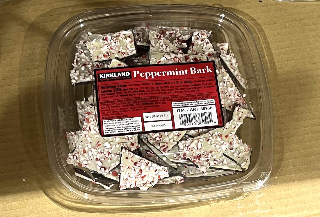 A clear plastic container of Kirkland brand Peppermint Bark sits on a cardboard surface, showing pieces of white chocolate bark topped with crushed peppermint candy inside. A red and black label is visible on the lid.