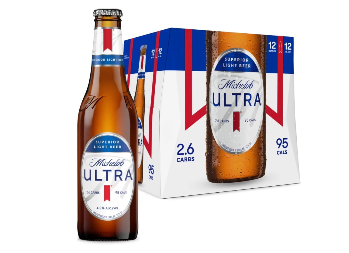 A Michelob Ultra beer bottle stands next to a 12-pack box. The bottle and box display the Michelob Ultra logo, blue and red accents, and highlight “2.6 carbs” and “95 calories” per serving.