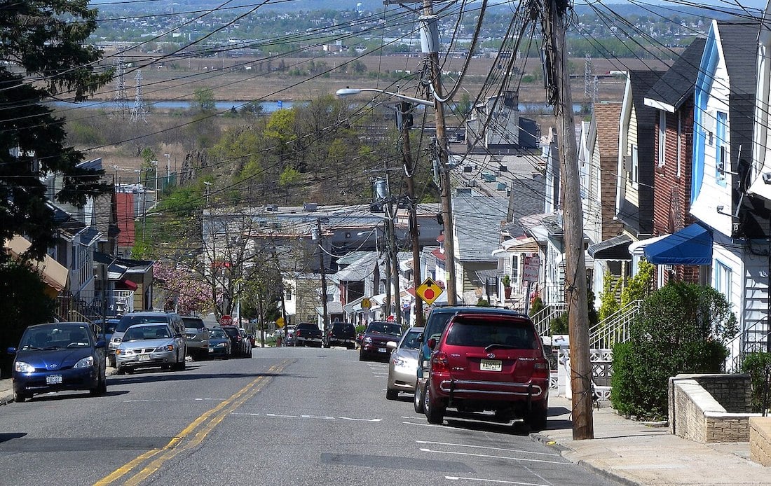 A suburban street on a hill with parked cars on both sides, houses with porches, trees, and many overhead utility wires. In the distance, there are fields, water, and a hazy skyline.