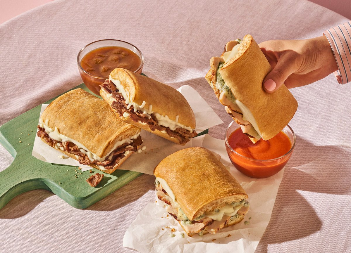 Four toasted sandwiches from the new fast-food menu, filled with meat and melted cheese on ciabatta bread, sit on parchment paper. A hand picks one up, while two bowls of red dipping sauce rest nearby on a pink tablecloth.