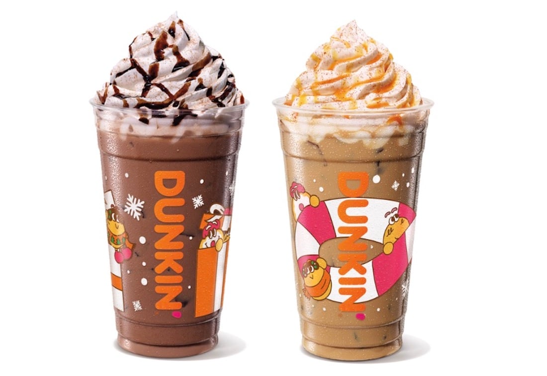 Two Dunkin' drinks in clear cups with holiday-themed designs. The left drink is chocolate with whipped cream and chocolate drizzle; the right is light brown with whipped cream and cinnamon.