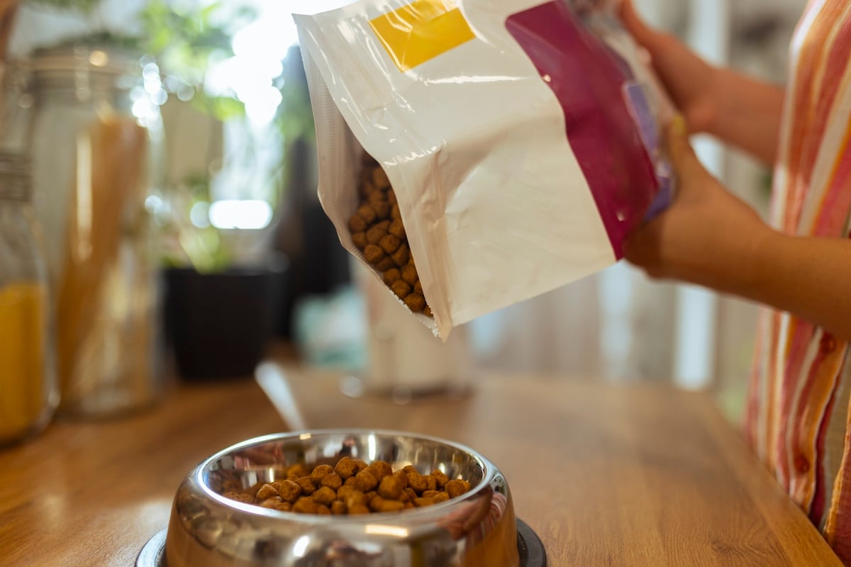 A person pours dry pet food from a bag into a stainless steel bowl on a wooden table, with kitchen items visible in the background.