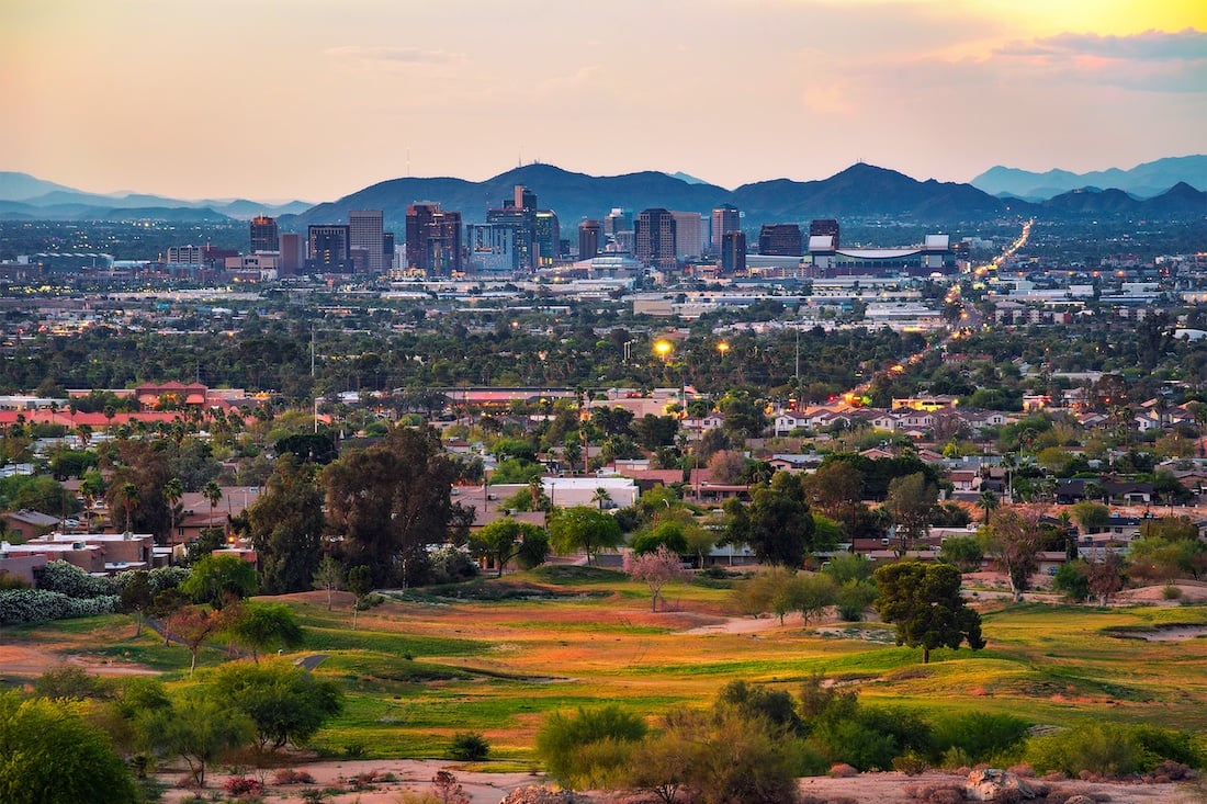 A view of Phoenix, Arizona at sunset, showing a mix of residential areas, green spaces, and the downtown skyline with mountains in the background under a partly cloudy sky.