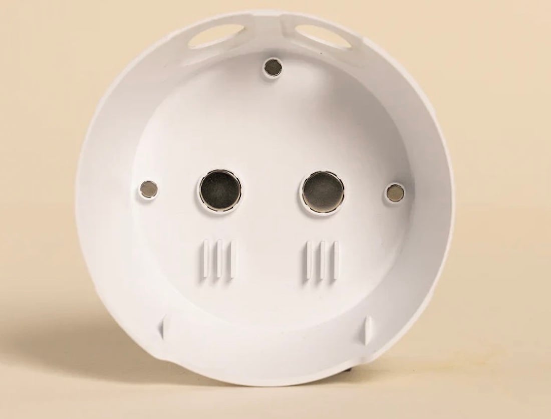 A white, round plastic electrical junction box is shown against a beige background, with two metal screw holes and two smaller mounting holes visible inside.