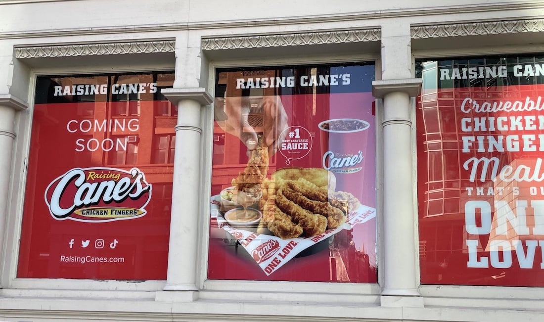 Red storefront window signs announce a "Raising Cane's Chicken Fingers" restaurant coming soon. The center features a meal with chicken fingers, fries, sauce, and a drink.