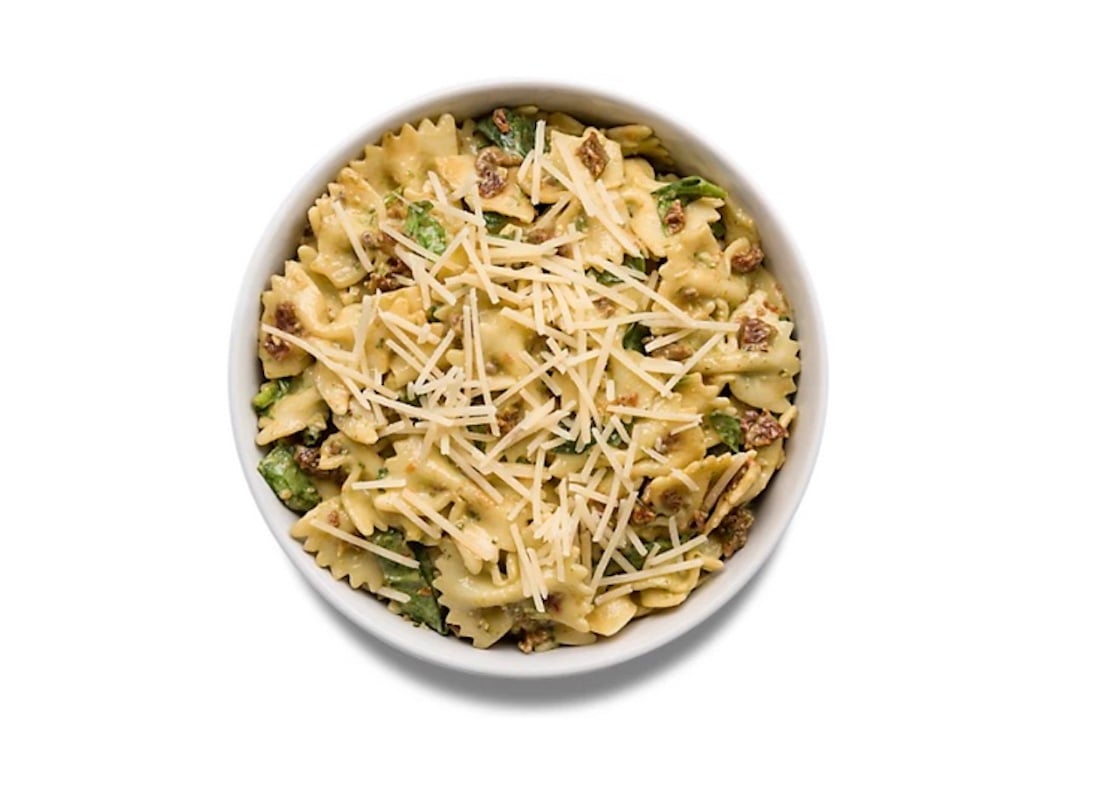 A white bowl filled with bow-tie pasta mixed with leafy greens, ground meat, and topped with shredded cheese, viewed from above on a white background.
