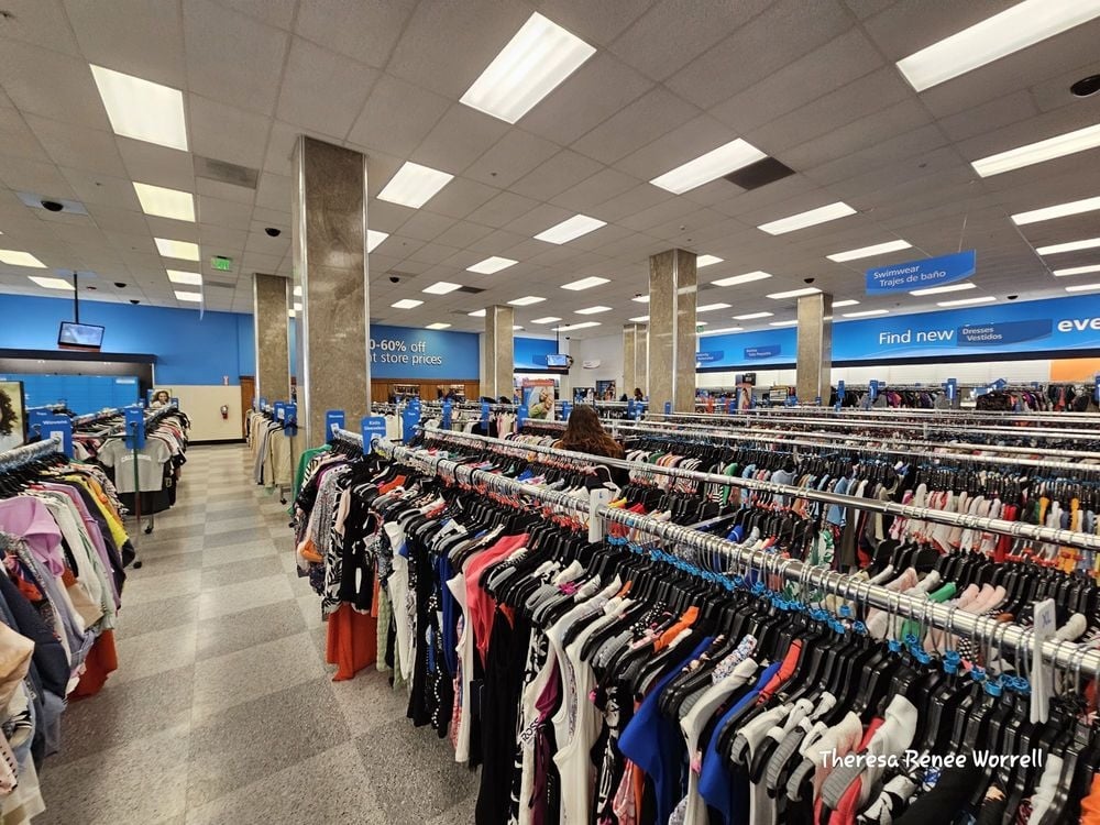 Clothing racks filled with colorful clothes inside a large, well-lit thrift store, with shoppers browsing and blue sale signs visible throughout the store—it's a scene you'd find at Ross Stores or your local DDS Discounts.