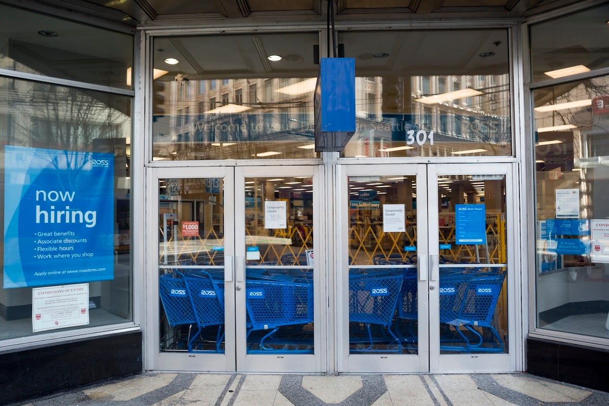 Storefront entrance with glass doors, blue shopping carts inside, and a large "now hiring" sign listing job benefits on the left window. The store appears to be welcoming applicants.