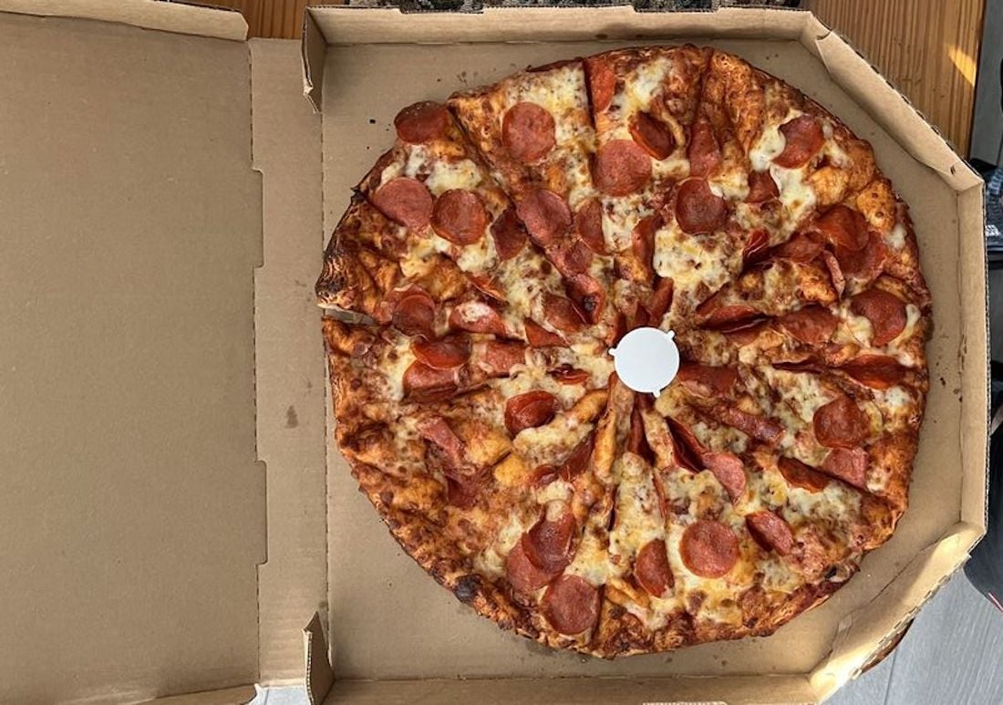 A pepperoni pizza cut into many irregular slices sits in an open cardboard pizza box, viewed from above. A small plastic pizza saver is placed in the center.