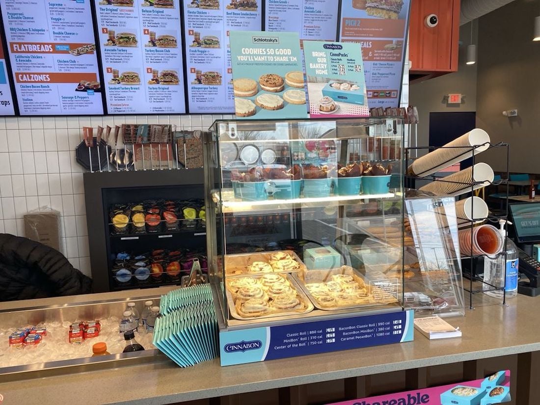 A bakery counter at Cinnabon displays trays of cinnamon rolls, cookies, and other treats. Menus hang above the counter, and there are stacks of napkins, condiments, and disposable drink holders on the counter.