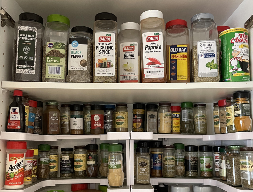 A kitchen cabinet filled with neatly organized spice containers of various sizes and brands, arranged in rows on white shelves. Labels such as paprika, oregano, black pepper, and pickling spice are visible.