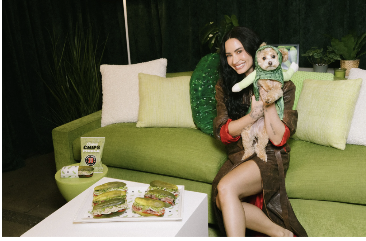 A woman sitting on a green couch holds a small dog dressed in a green costume. In front of her, there is a table with sandwiches and a bag of chips. The setting is cozy with green and white pillows and plants.