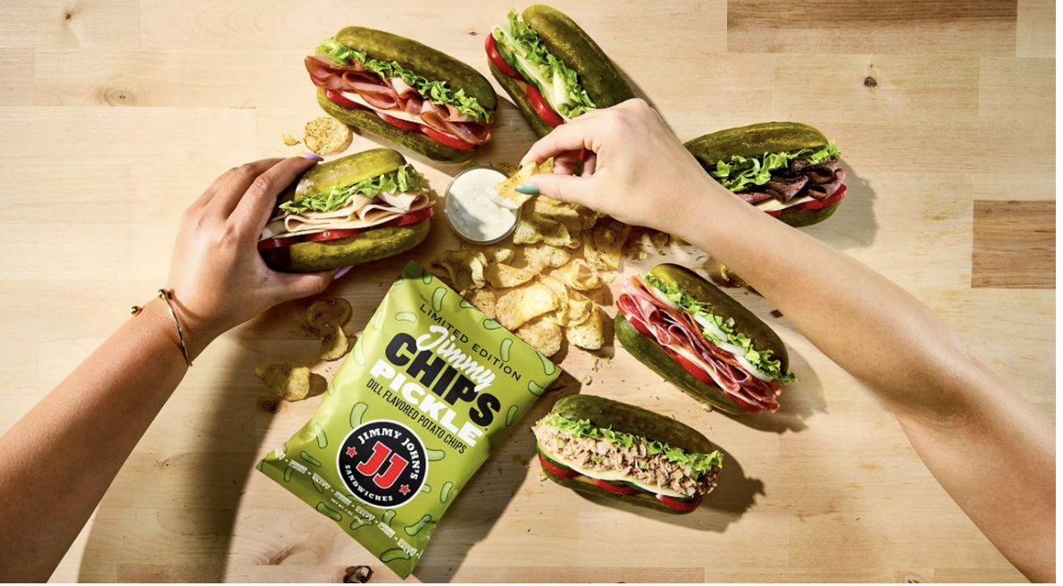Two hands reach for a spread of pickle sandwiches filled with meats and veggies, surrounded by potato chips and a bag of Jimmy John’s chips on a wooden table. One hand dips a chip into a white sauce.