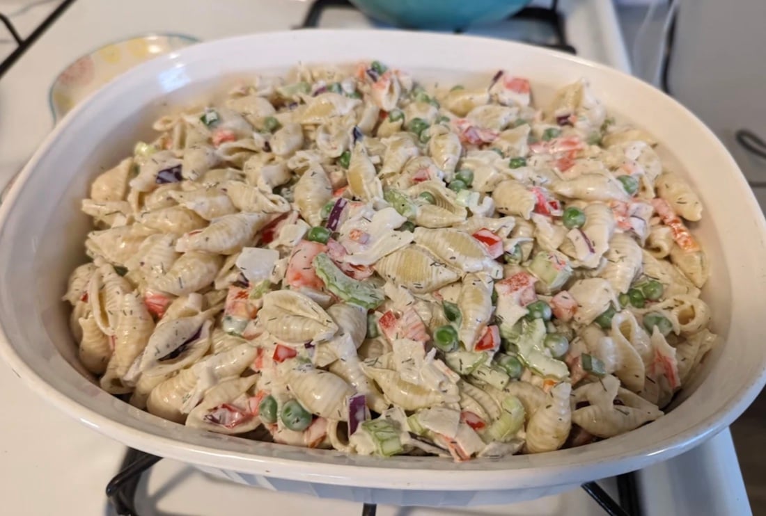 A casserole dish filled with pasta shells mixed with peas, diced red peppers, celery, and a creamy dressing, sitting on a stovetop.