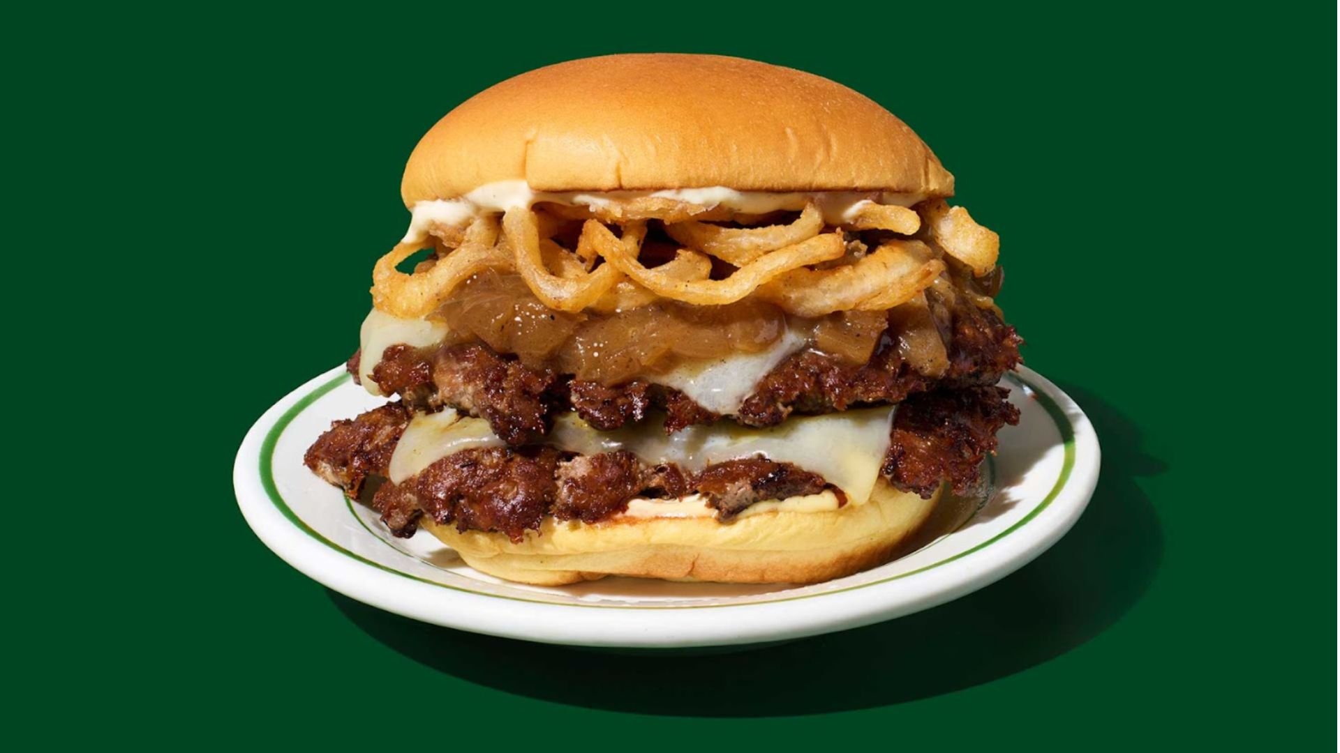 A double cheeseburger from a new fast-food menu sits on a white plate, topped with crispy fried onions, melted cheese, caramelized onions, and a toasted bun, against a solid green background.