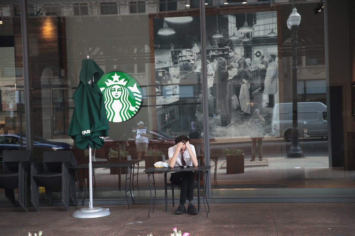 A person sits alone at an outdoor table in front of a Starbucks, resting their head in their hands. Empty chairs and a folded green umbrella are nearby, with a large Starbucks logo and vintage photo on the window behind them.