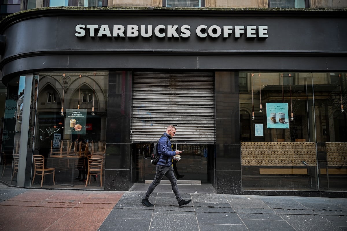 A man walks past a closed Starbucks Coffee shop with a metal shutter partially down, empty chairs visible inside, and posters on the windows.