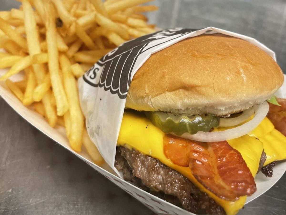 A close-up of a cheeseburger with bacon, pickles, onions, and melted cheese in a wrapper from fast-food chains that never use a microwave, served with thin, crispy French fries in a paper tray.