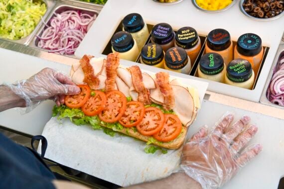 A person wearing gloves assembles a sub sandwich with lettuce, tomato slices, turkey, and bacon in front of sauces and toppings—perhaps creating one of the Subway secret menu items at the sandwich shop counter.