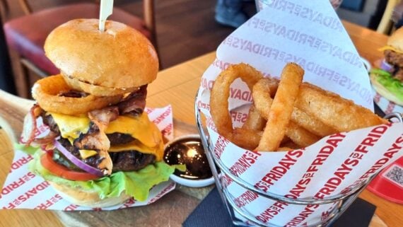 A cheeseburger with lettuce, tomato, red onion, bacon, and onion rings sits next to a metal cup filled with curly fries, both served on TGI Fridays-branded paper on a wooden table.