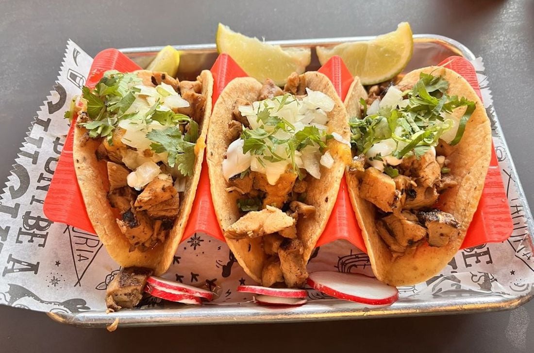 Three tacos filled with grilled chicken, topped with chopped onions and fresh cilantro, served on a tray with lime wedges and sliced radishes on the side.