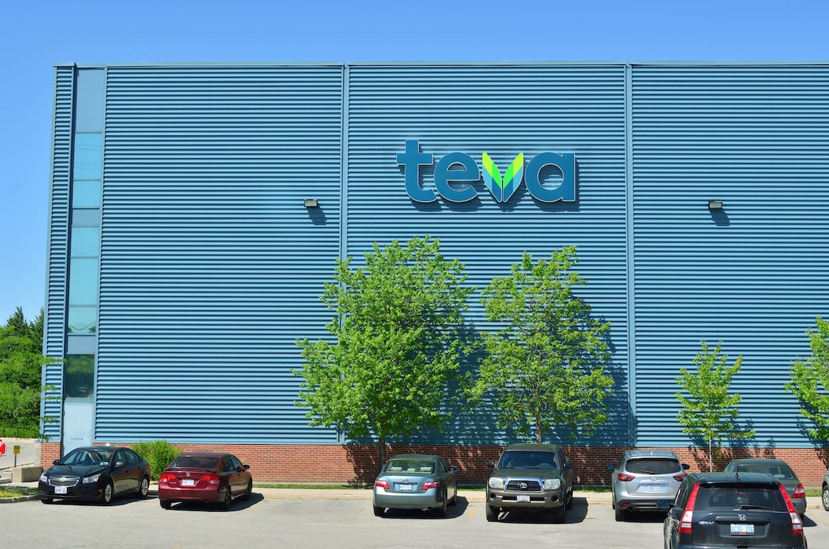 A large blue industrial building with the "teva" logo on the wall, several trees in front, and cars parked in the parking lot on a sunny day.