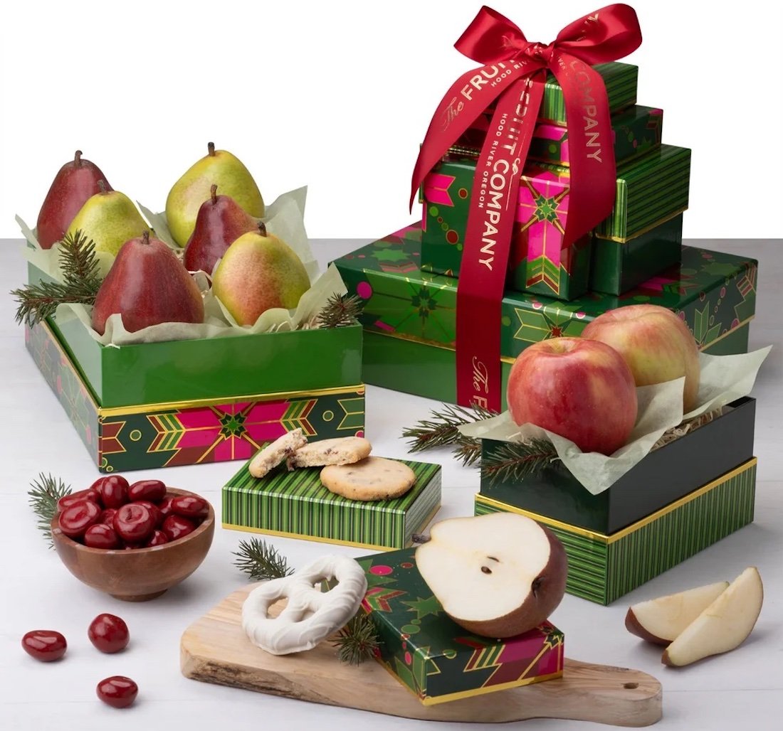 A festive gift set with green boxes containing pears, apples, cookies, pretzels, and cherries, some boxes tied with red ribbon. Fruits and snacks are arranged on a wooden board and around the boxes with sprigs of pine.