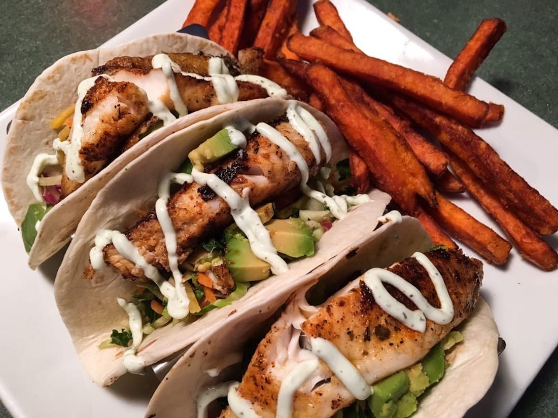 Three grilled fish tacos topped with avocado, slaw, and creamy sauce on flour tortillas, served with a side of sweet potato fries on a white plate.