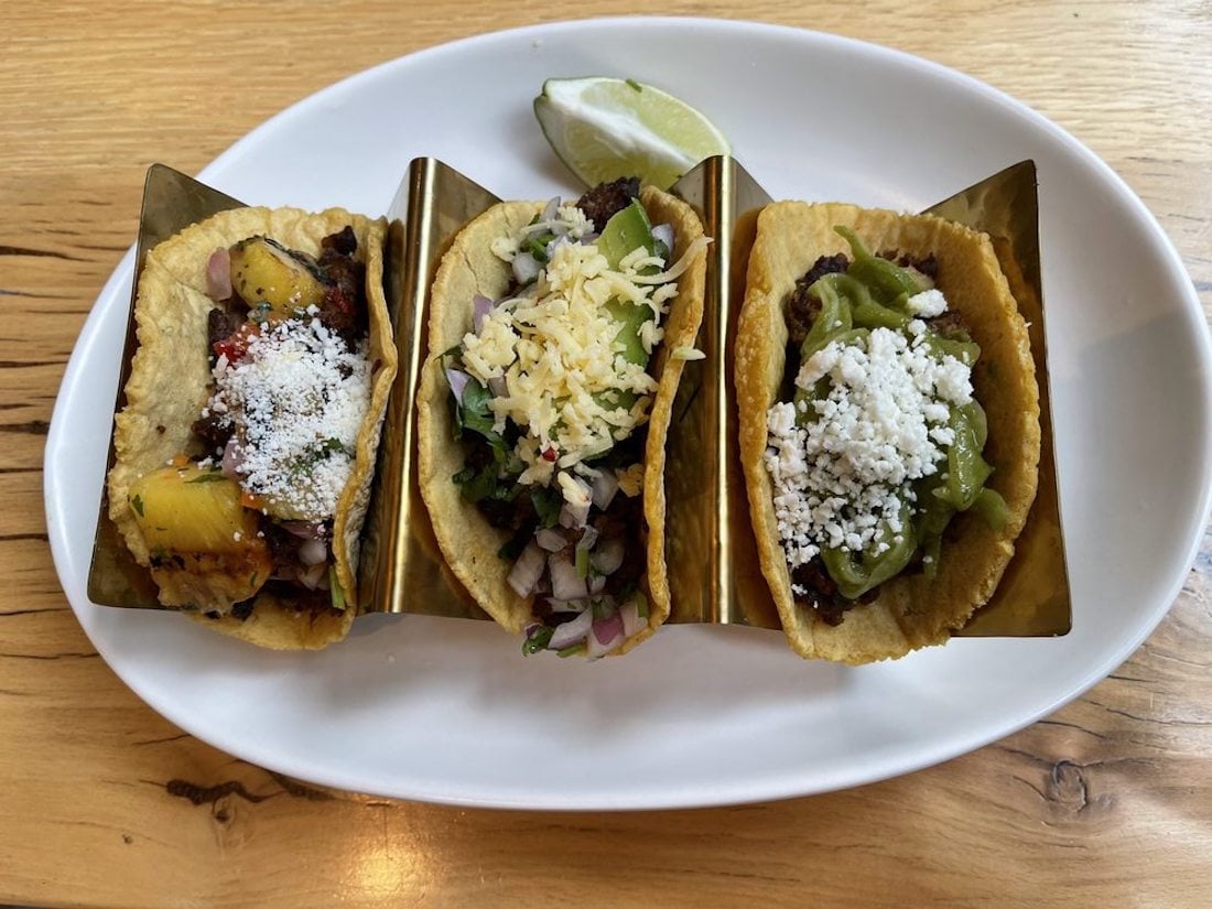 Three tacos in taco holders on a white plate, each topped with different ingredients such as cheese, guacamole, diced vegetables, and grilled pineapple, with a lime wedge in the background.