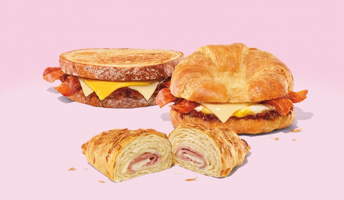 Three breakfast items on a pink background: a bacon and cheese sandwich on toasted bread, a bacon, egg, and cheese croissant, and a ham and cheese croissant, sliced to show the filling.