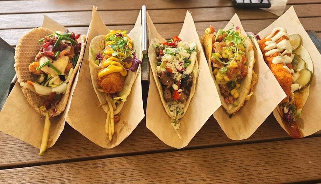 Five assorted tacos filled with various ingredients, such as fried fish, grilled meat, fresh vegetables, cheese, pickles, and colorful sauces, are arranged in brown paper holders on a wooden table.