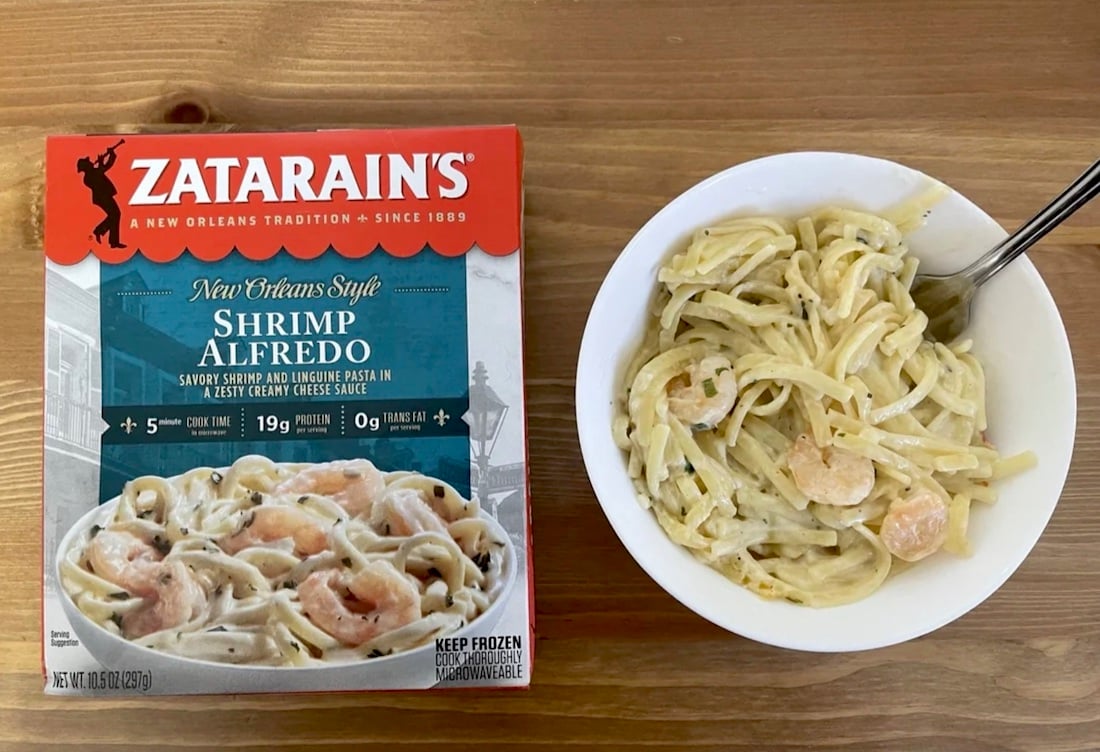 A box of Zatarain’s New Orleans Style Shrimp Alfredo frozen meal sits next to a white bowl filled with shrimp alfredo pasta on a wooden table. A fork is in the bowl.