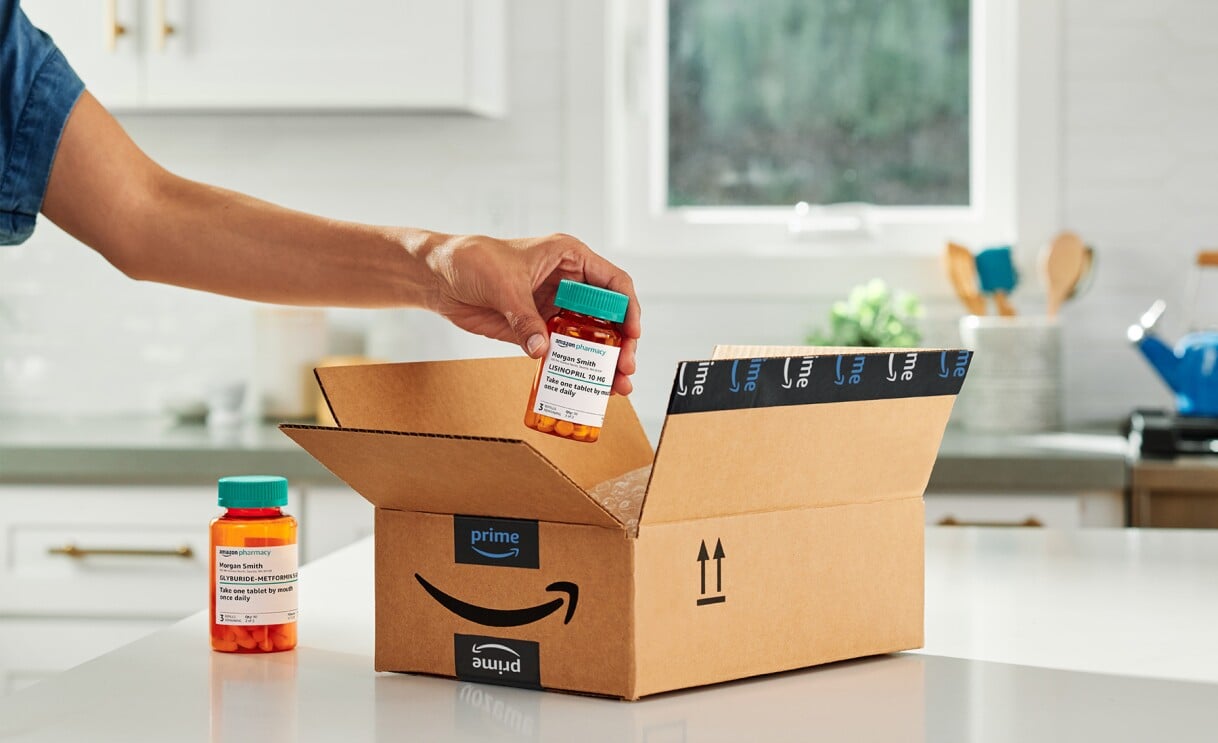A person’s hand places a prescription pill bottle into an open Amazon Prime box on a white kitchen counter, with another pill bottle beside the box and a bright window in the background.