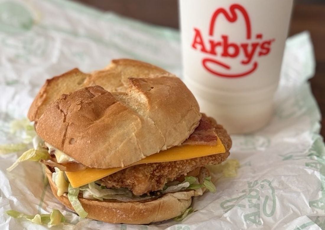 A crispy chicken sandwich with lettuce, cheddar cheese, and bacon on a bun, served on Arby’s-branded paper next to an Arby’s soft drink cup.