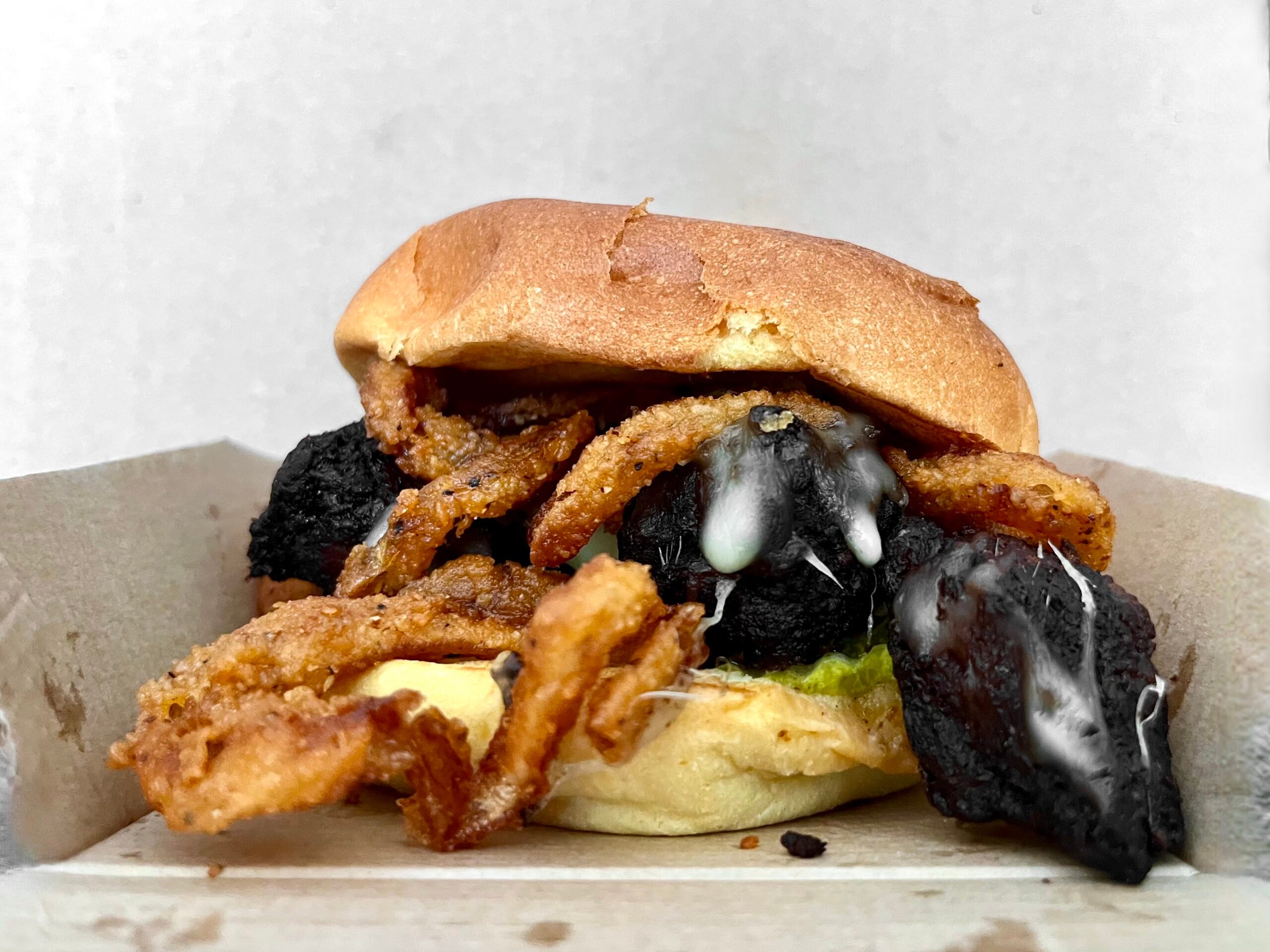 A close-up of a sandwich in a cardboard tray, filled with crispy fried onions, melted cheese, charred or blackened pieces, and a soft bun, against a plain background.