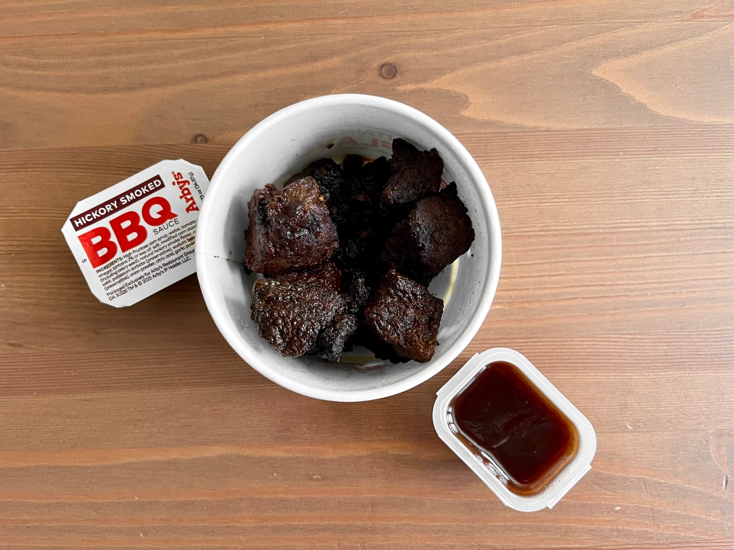 A white bowl filled with several pieces of dark, cooked meat sits on a wooden table next to two containers of Hickory Smoked BBQ sauce, one opened and one closed.
