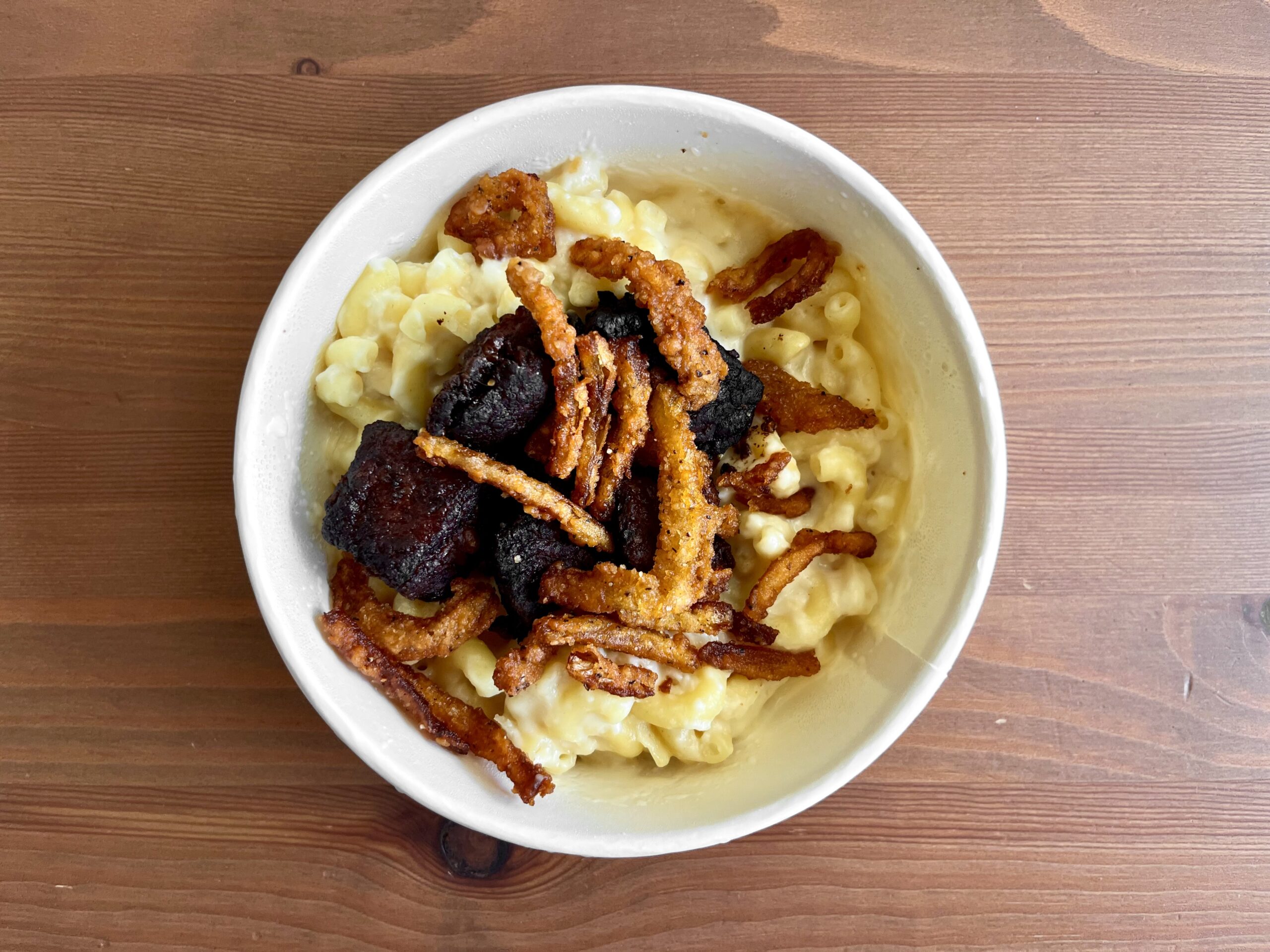 A bowl of macaroni and cheese topped with crispy fried onions and chunks of dark, browned meat, placed on a wooden surface.