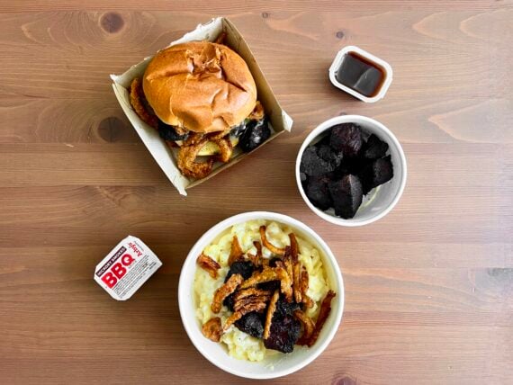 Overhead view of a BBQ meal on a wooden table with a pulled pork sandwich, a bowl of macaroni and cheese topped with BBQ meat, a bowl of burnt ends, and two containers of BBQ sauce.
