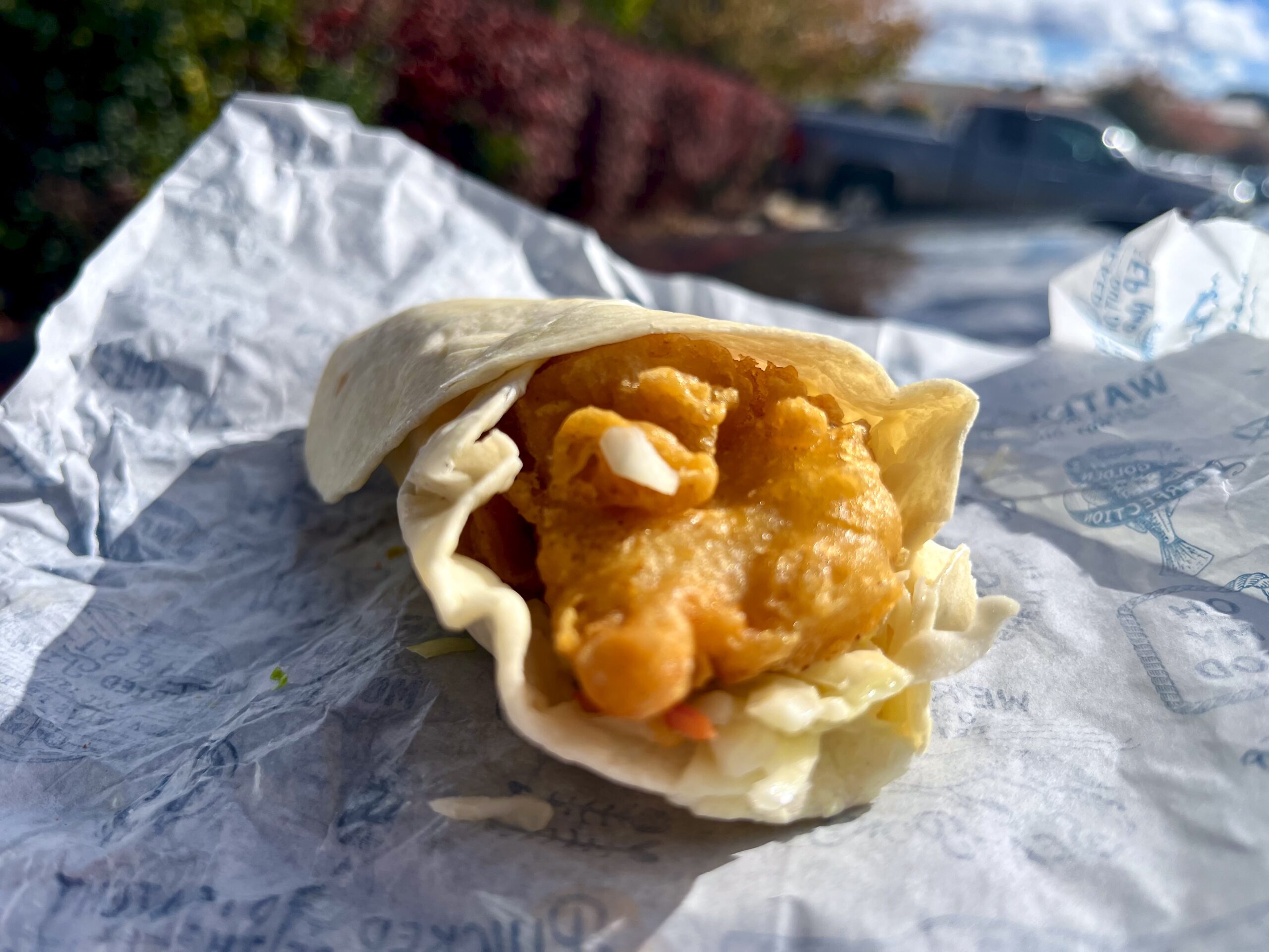 A close-up of a soft tortilla wrap filled with crispy fried chicken and shredded lettuce, resting on crumpled white paper, with an outdoor background visible.