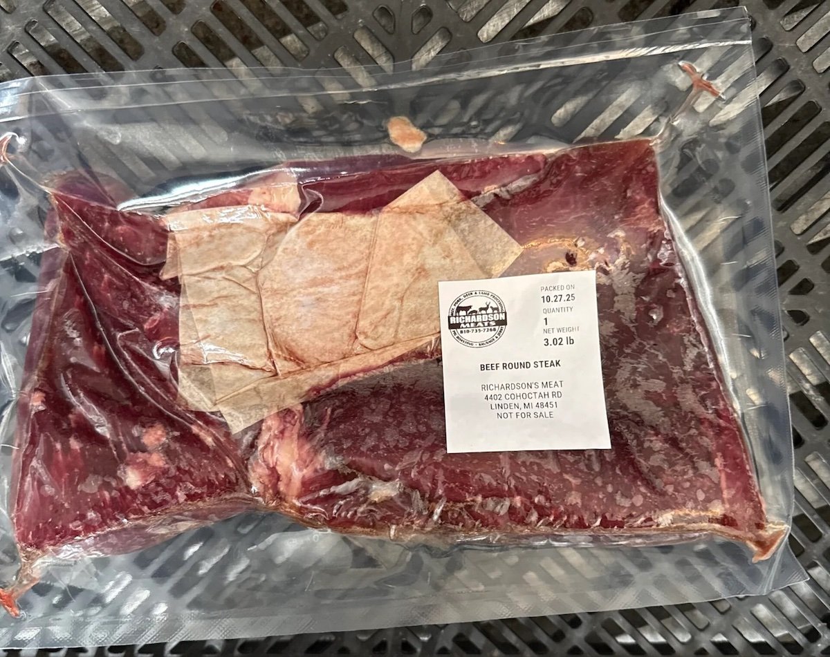 A vacuum-sealed package of beef round steak sits on a black plastic surface. The label shows product details, including weight (3.02 lbs), cut type, and farm information, with a visible USDA stamp.