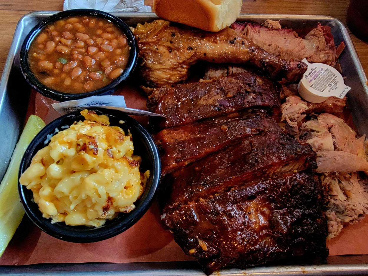 A tray of the best BBQ food with ribs, pulled pork, brisket, a chicken leg, baked beans, macaroni and cheese, a pickle spear, a slice of bread, and a portion of butter in a small cup.