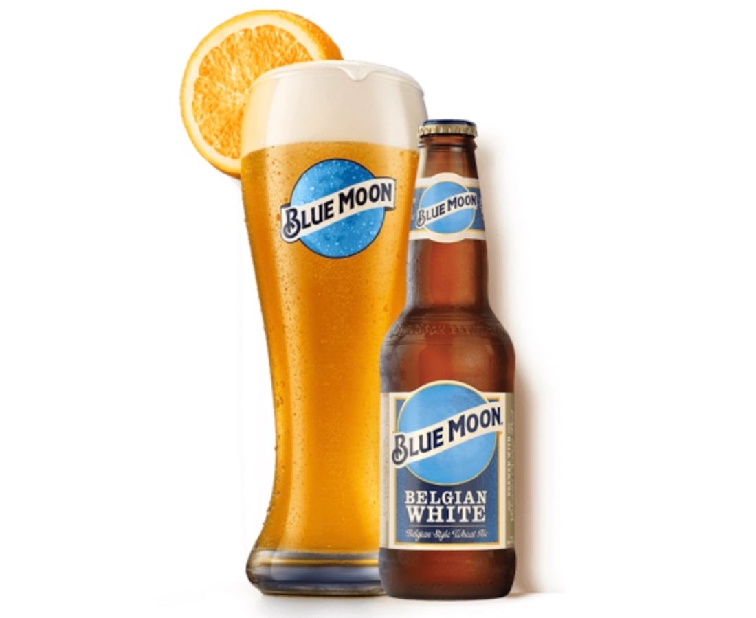 A bottle of Blue Moon Belgian White beer stands next to a tall glass of the beer garnished with an orange slice. Both the bottle and glass display the Blue Moon logo.