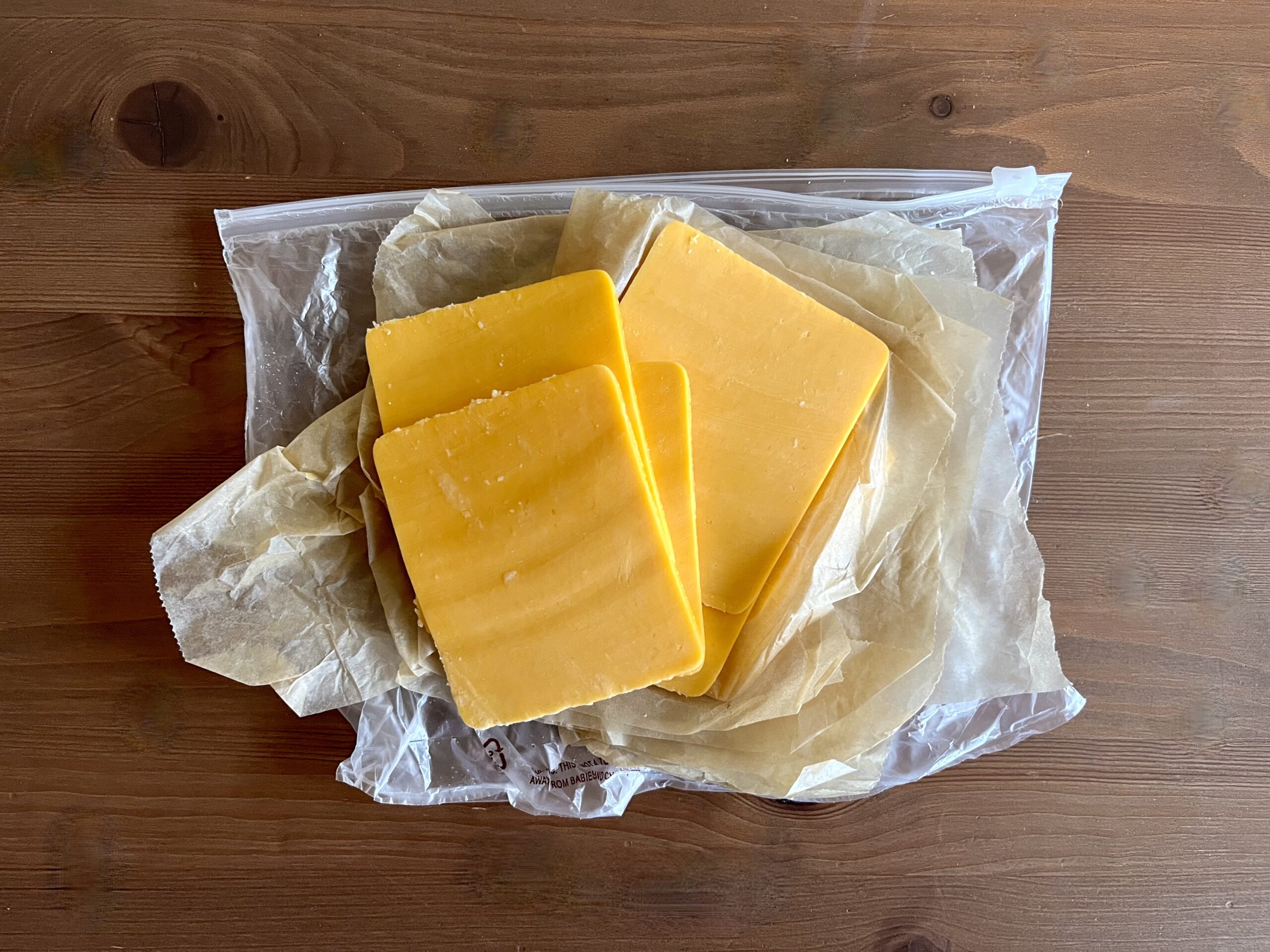 Three slices of boars head deli-sliced cheddar cheese sit on brown wax paper inside a resealable plastic bag, placed on a wooden surface.