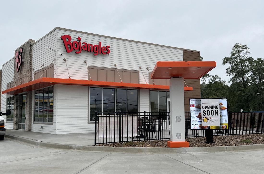 A Bojangles restaurant with a drive-thru menu sign in front displaying "Opening Soon." The building is white with orange accents, and the sky is overcast.