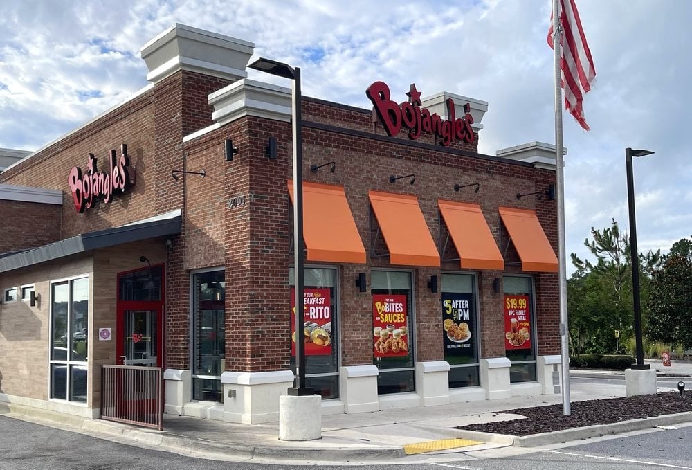 A Bojangles restaurant with a brick exterior, orange awnings, and large menu posters in the windows. An American flag is raised on a pole outside. The sky is partly cloudy.